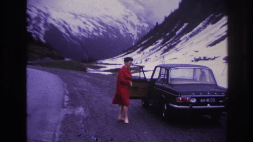 8mm film still, France c.1967 — woman in red dress standing beside the open rear door of a dark European saloon on a French mountain road, snow-patched slopes rising steeply behind.