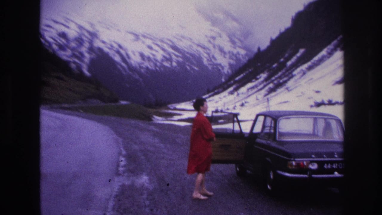 Woman in red dress next to a dark European sedan on a narrow alpine road, possibly France circa 1967; snow-covered mountain peaks visible in background.