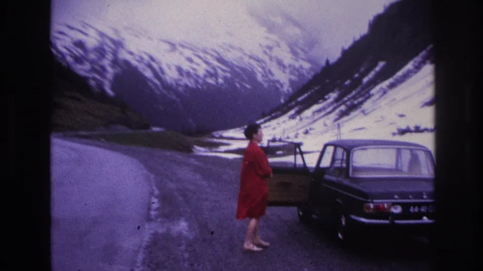 Woman in red dress next to a dark European sedan on a narrow alpine road, possibly France circa 1967; snow-covered mountain peaks visible in background.