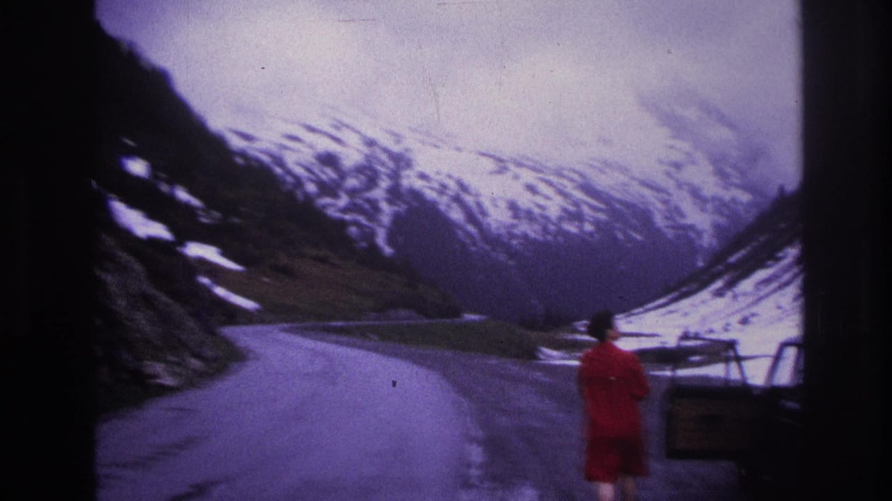 Woman in red seen from behind on a mountain road; dark sedan at right frame edge; snow-covered alpine peaks in background.