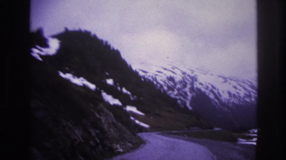Panning shot of a snow-patched alpine valley with a narrow winding road and rocky crags, filmed on 8mm, possibly French Alps circa 1967.