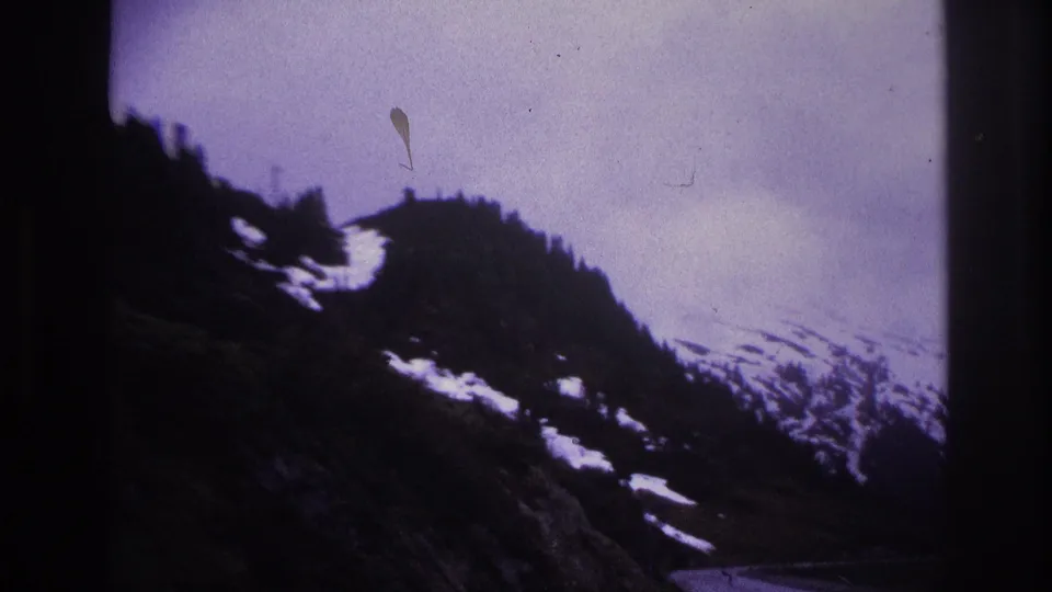 End-of-pan frame showing a rocky alpine ridgeline with snow patches, sparse conifers, and a visible film-emulsion artifact; likely French Alps, c. 1967.