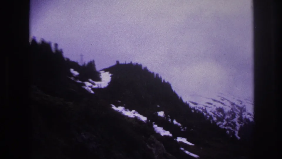 Aged 8mm film frame showing snow-covered alpine peaks under a moody overcast sky, likely French Alps, 1967.