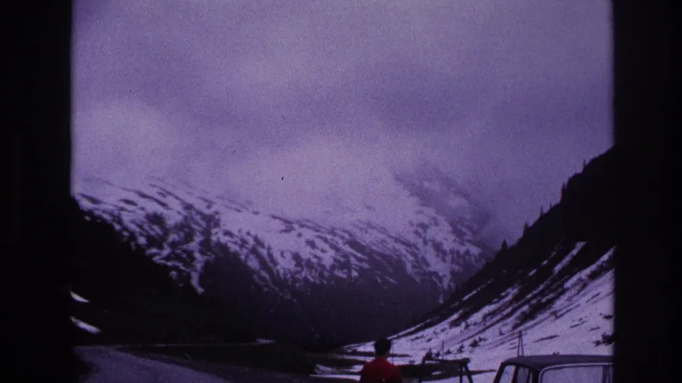 8mm home movie showing a broad alpine valley; a person in a red jacket stands beside a parked car at a roadside stop, France, 1967.