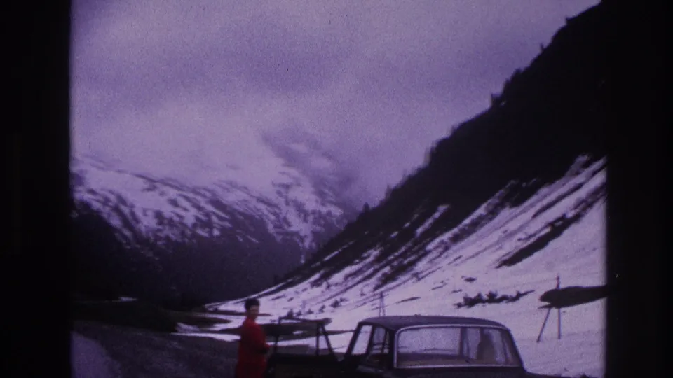 8mm home movie of a dramatic alpine valley with a person in a red jacket and a parked classic car at a roadside stop, likely French Alps, 1967.