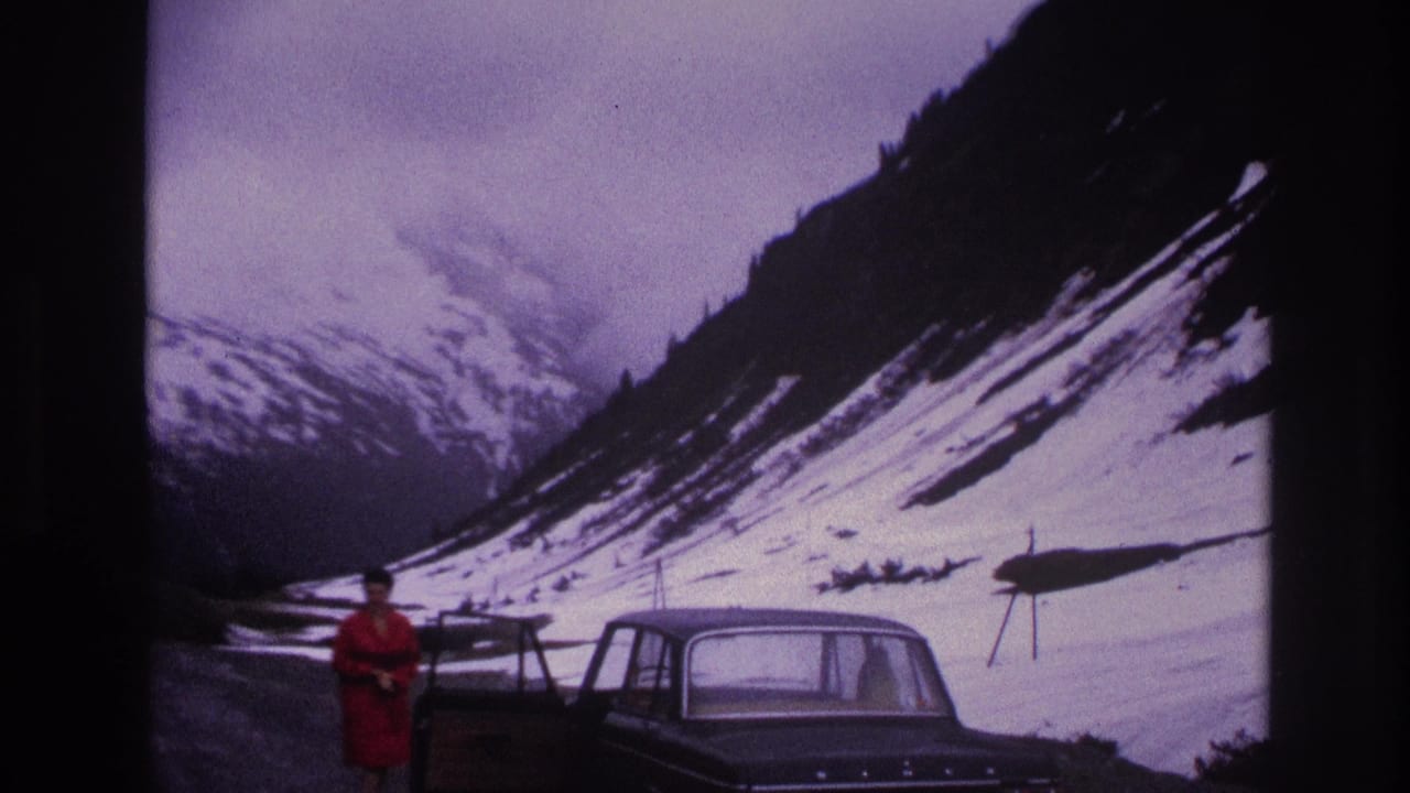 Woman in red coat beside a dark 1960s sedan on a gravel mountain road in France, circa 1967; snow-covered Alpine slopes and overcast sky in background.