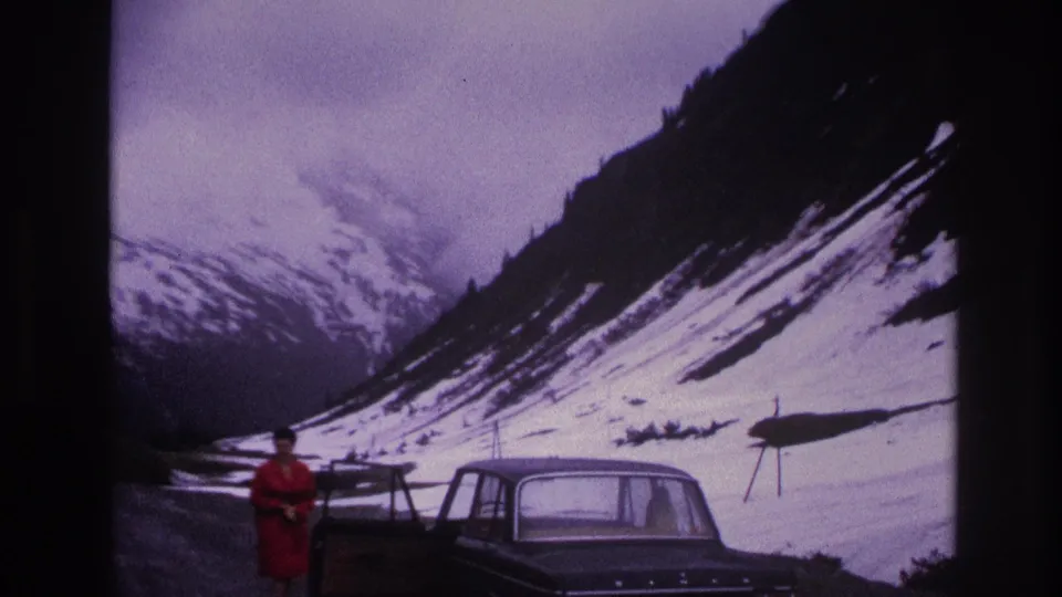 Woman in red coat beside a dark 1960s sedan on a gravel mountain road in France, circa 1967; snow-covered Alpine slopes and overcast sky in background.