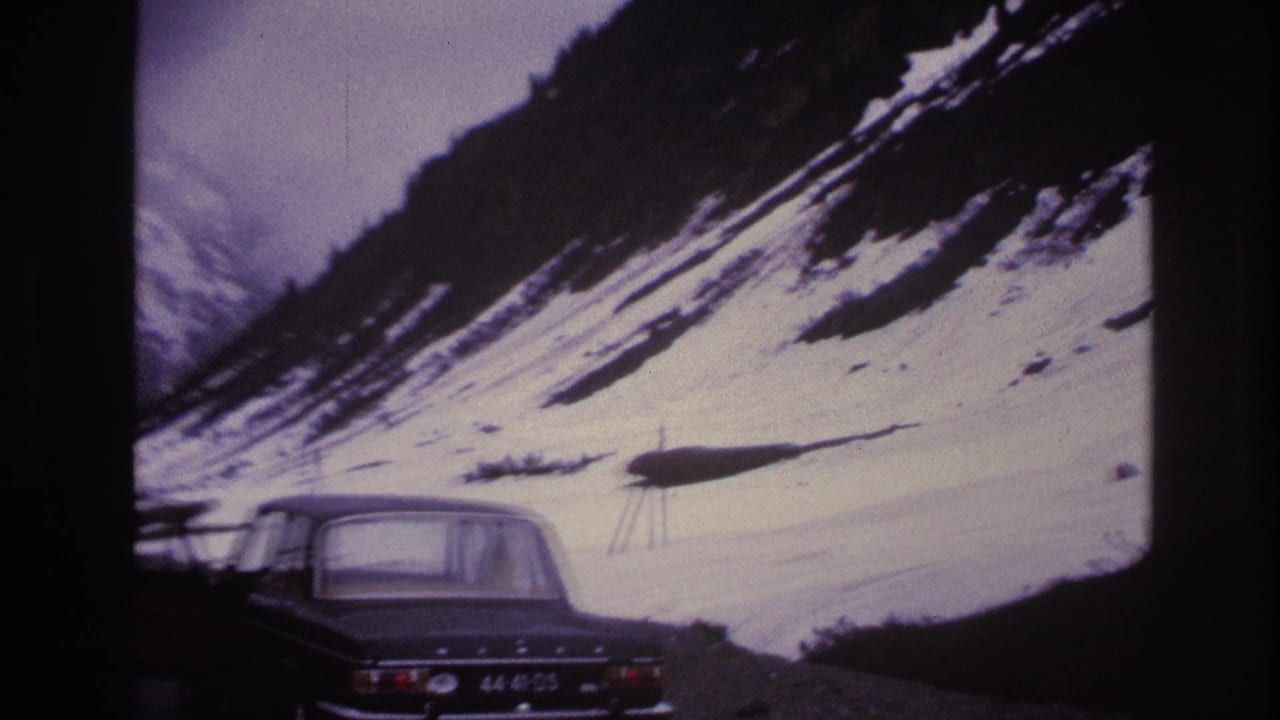 Rear view of a dark 1960s sedan on a gravel mountain road, license plate legible, snow-covered Alpine slope and pylons visible behind.