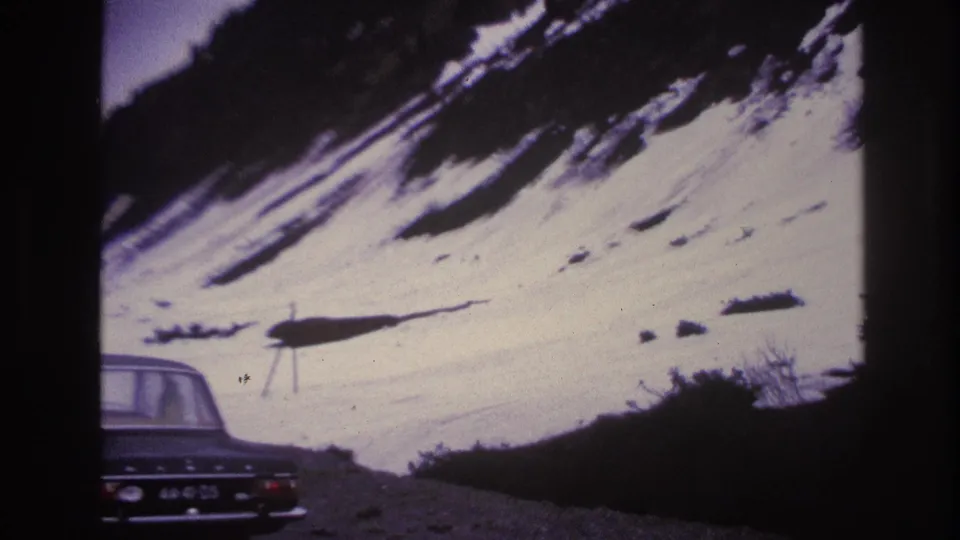 Wide view of a steep snow-covered Alpine slope in France, circa 1967; a dark 1960s sedan at lower left with legible license plate, ski lift pylons visible on the hillside above.