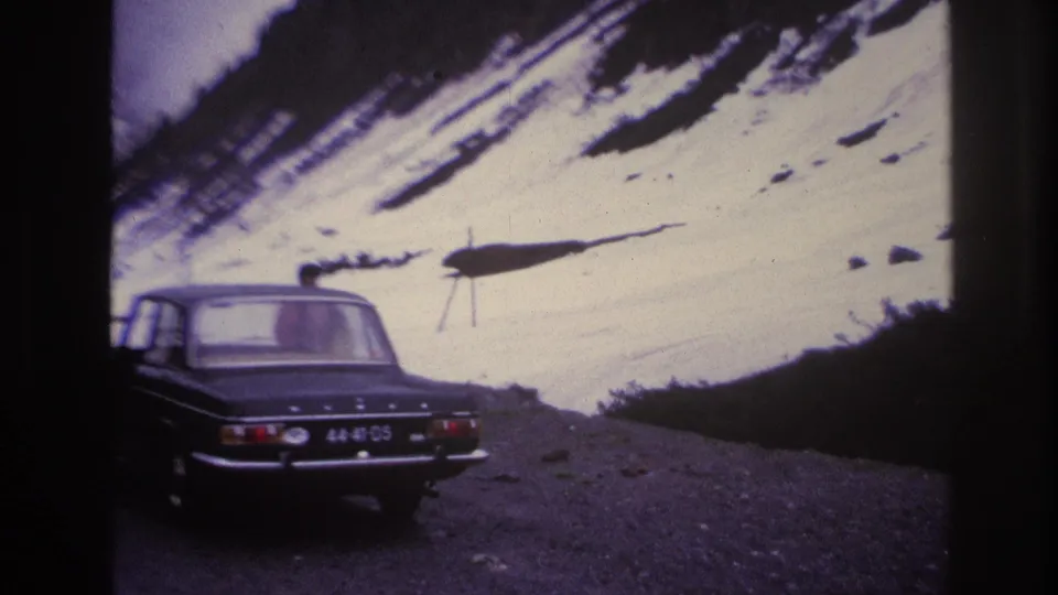 Dark 1960s sedan on a French Alpine gravel road, license plate characters visible; steep snow-covered slopes and apparent ski lift structure in background, circa 1967.