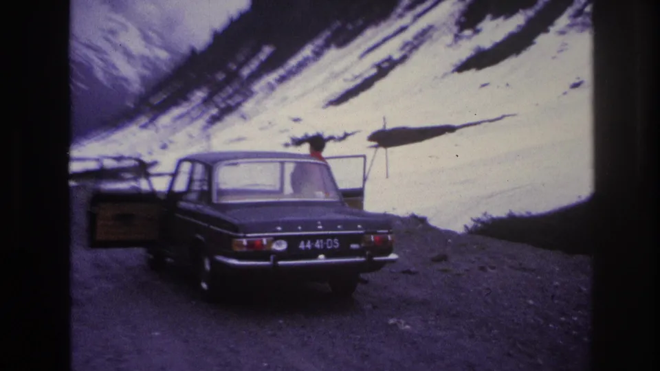 Woman in red coat entering the rear passenger door of a dark European sedan on a French mountain road, c.1967; a second vehicle or luggage crate visible at left edge.