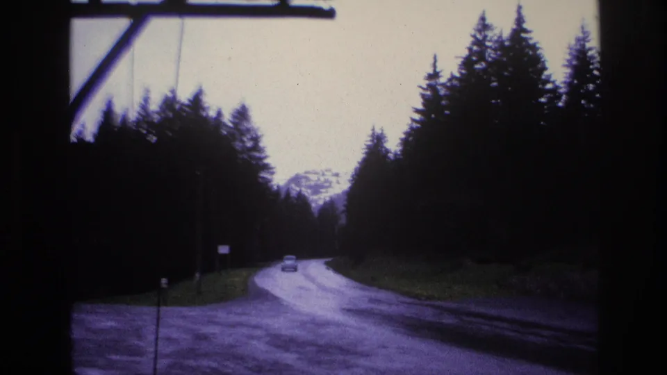 Winding alpine road flanked by conifer trees with mountain peaks in background, a small distant vehicle on the road; consistent with French Alps, c.1967. A long dark object crosses the upper frame edge.