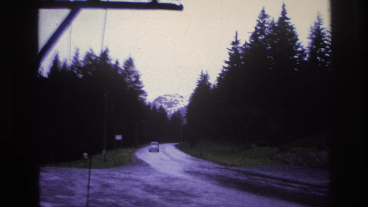 8mm shot from inside a moving car on a wet mountain road through dense evergreen forest, France circa 1967, with a snow-capped peak visible in the distance.