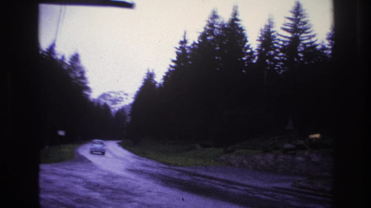 8mm footage from a moving vehicle on a winding French mountain road circa 1967, with a period compact car and alpine peak visible in the distance.