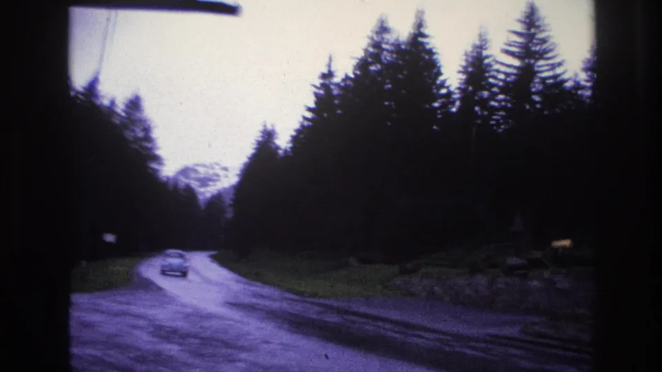 8mm footage from a moving vehicle on a winding French mountain road circa 1967, with a period compact car and alpine peak visible in the distance.