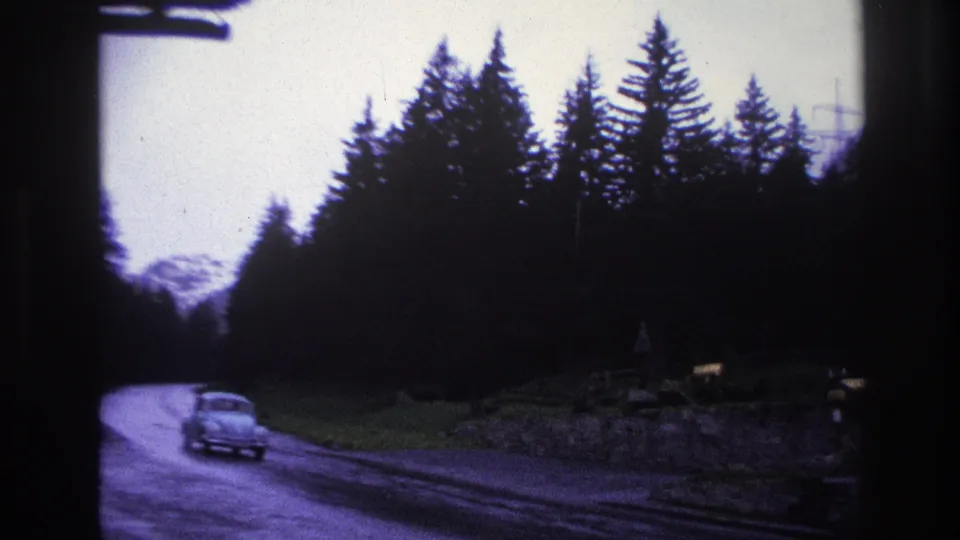 8mm footage of a rounded compact car on a wet mountain road through alpine forest, France circa 1967, with a snow-capped peak and utility poles visible.