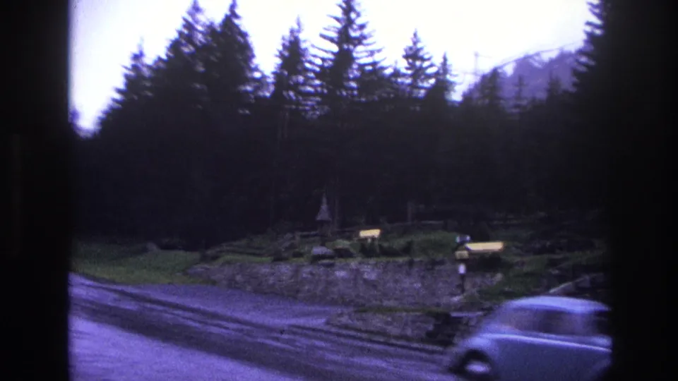 8mm footage of a vehicle passing close to camera on a French alpine mountain road circa 1967, with a small roadside structure and snow-capped peak in the background.