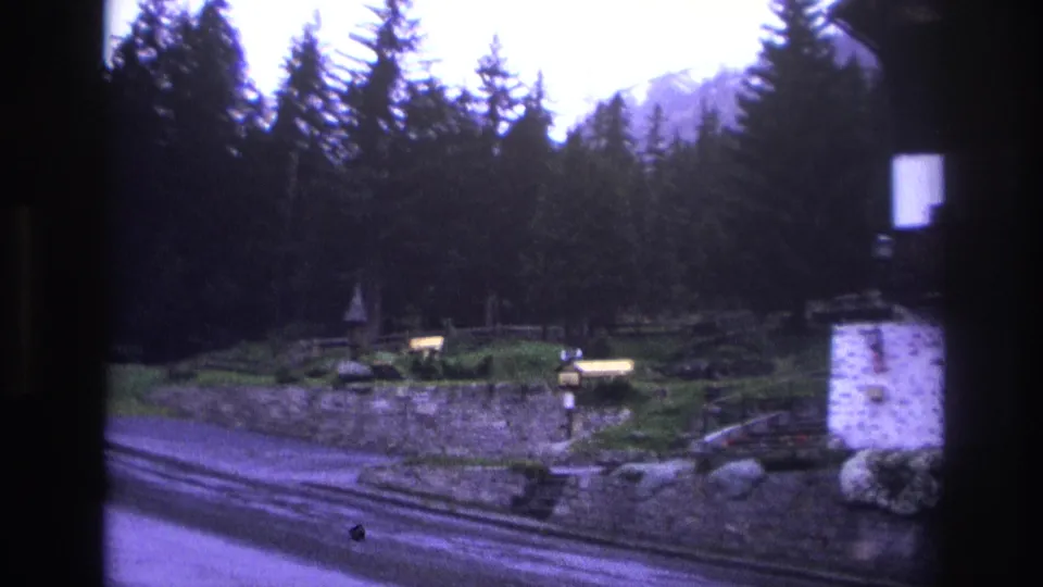 Mountain road with stone retaining wall and tall conifers, filmed in a French alpine setting circa 1967 on 8mm; strong purple color cast.