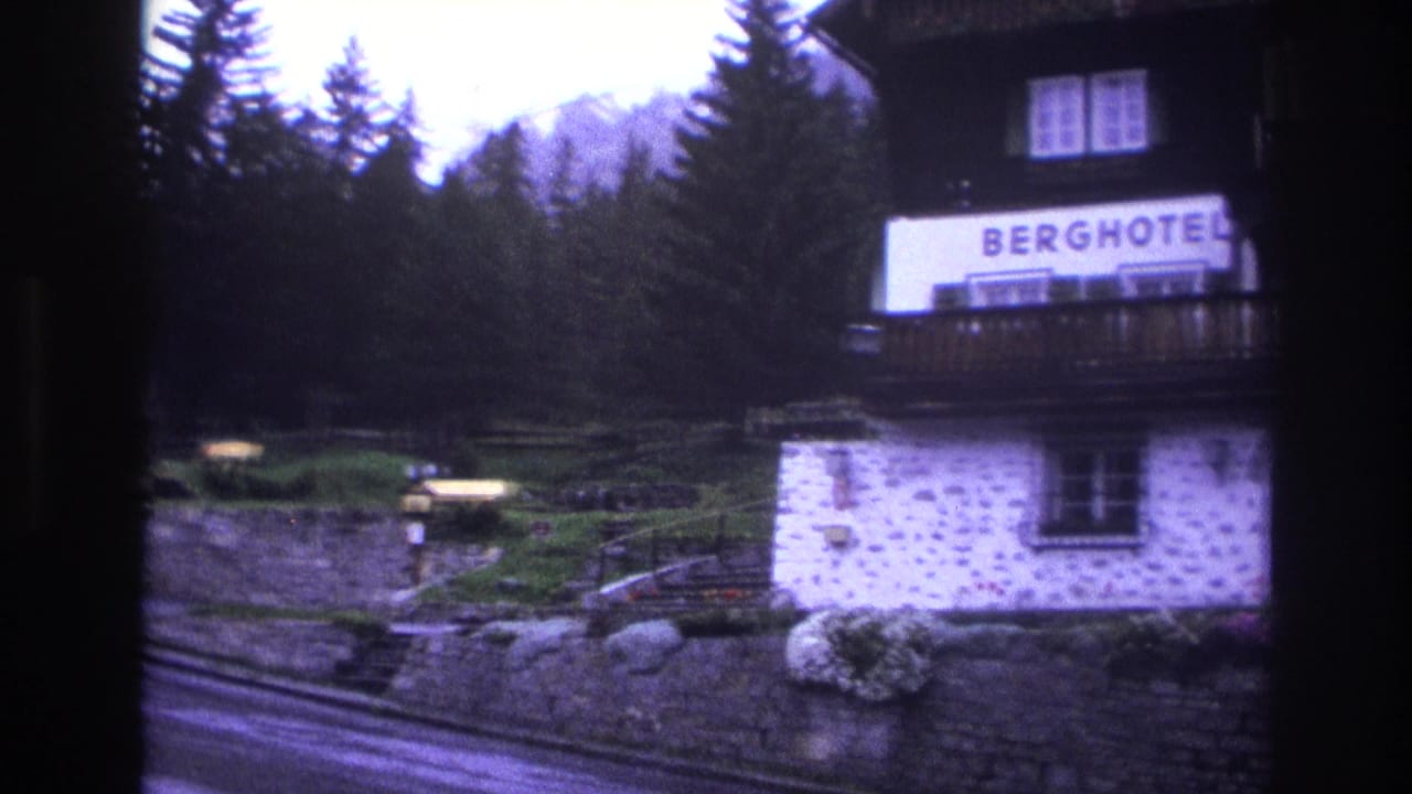 Berghotel building coming into view with legible BERGHOTEL sign, dark wood balcony, stone lower walls, conifers behind.