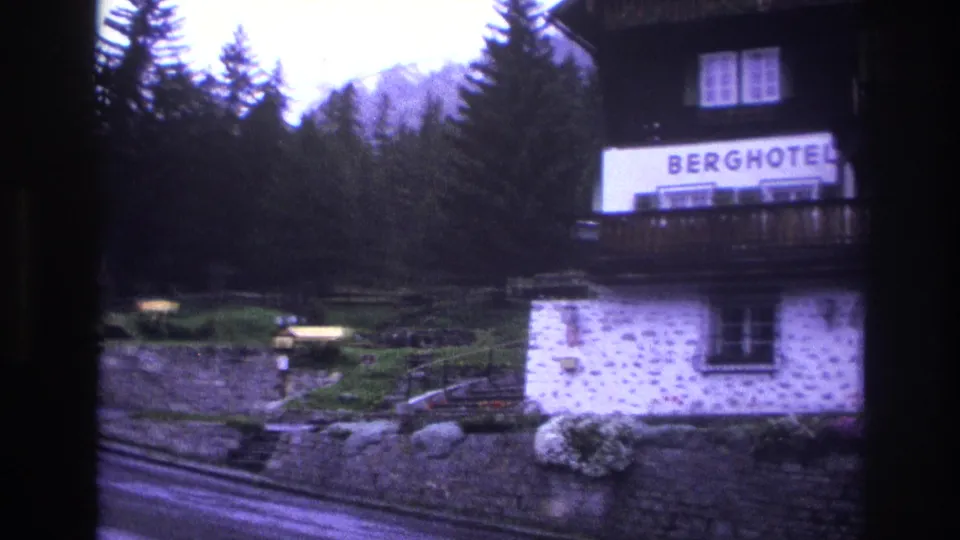 Alpine Berghotel with legible BERGHOTEL signage and dark wood balcony over stone masonry walls, filmed in France circa 1967 on 8mm with purple color cast.