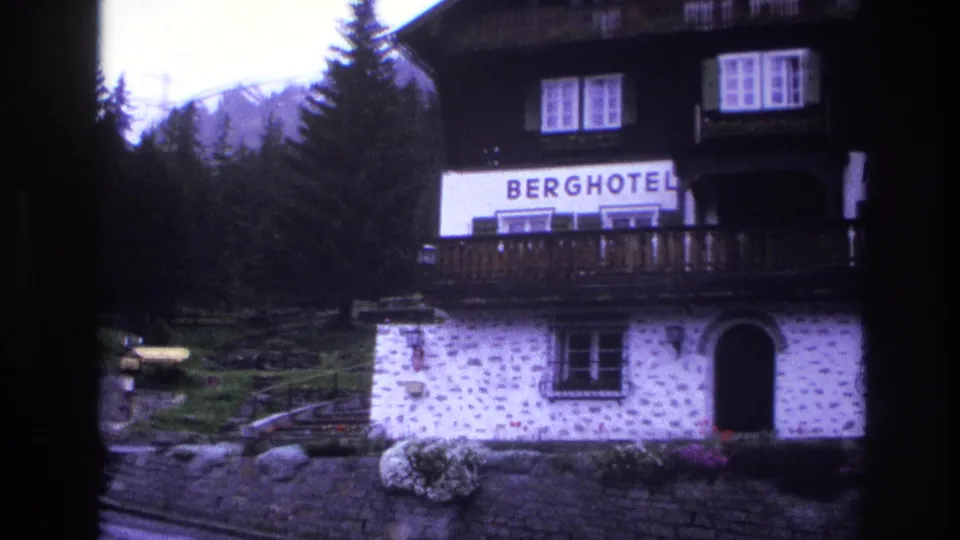 Full front facade of alpine Berghotel in France circa 1967: arched stone entrance, dark wood balcony, BERGHOTEL signage, flower planters, conifer forest and mountain peaks behind.