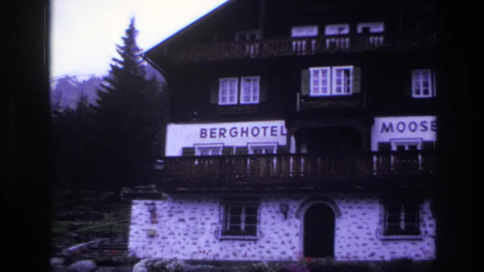 Upper facade of Berghotel Mooserkreuz, c.1967, with right side of sign partially cut from frame; dark timber chalet structure over white stonework; purple cast from aged 8mm stock.