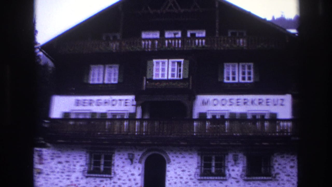 Full facade of Berghotel Mooserkreuz with complete sign banners, dark timber upper floors, white stone lower facade and evergreen trees behind.