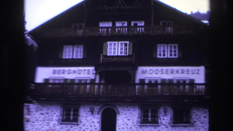 Full facade of Berghotel Mooserkreuz alpine hotel, c.1967; complete 'BERGHOTEL MOOSERKREUZ' sign banners legible, dark timber chalet above white stonework; purple 8mm color cast.