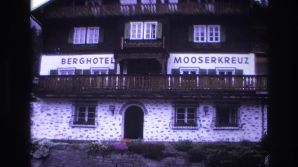Berghotel Mooserkreuz mid-to-lower facade, c.1967; sign banners prominent, arched stone entrance and flower boxes at ground level visible; purple 8mm color cast throughout.