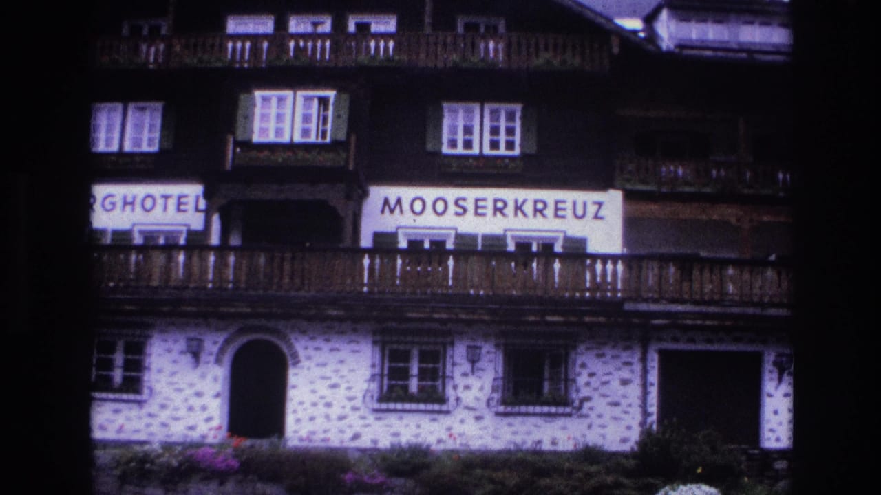 1967 French alpine hotel facade showing MOOSERKREUZ sign, wooden balconies, shuttered windows, whitewashed stone lower walls, and conifer hillside.