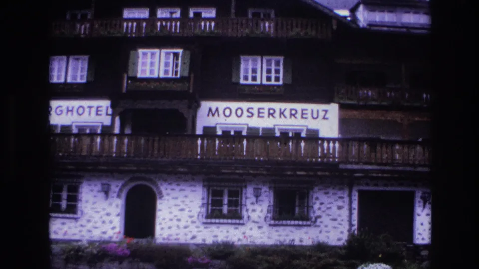 1967 French alpine hotel facade showing MOOSERKREUZ sign, wooden balconies, shuttered windows, whitewashed stone lower walls, and conifer hillside.