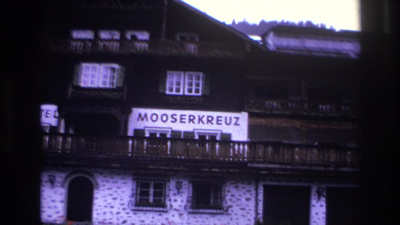 Closer view of Mooserkreuz hotel showing prominent sign, dark timber upper storey, stone lower walls, arched entry, and conifers at top right.