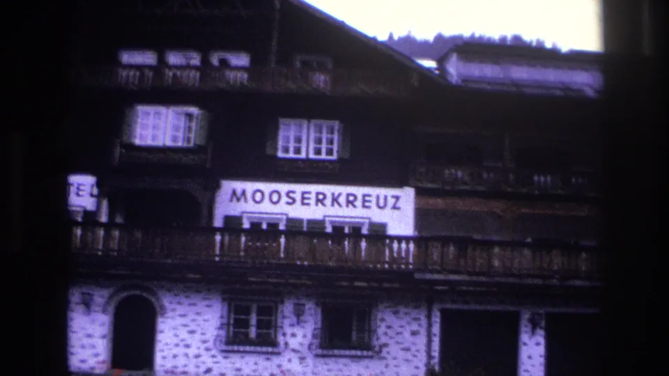 Mid-shot of Mooserkreuz alpine hotel, circa 1967 — sign clearly legible, dark timber balcony above whitewashed stone ground floor, conifers beyond.