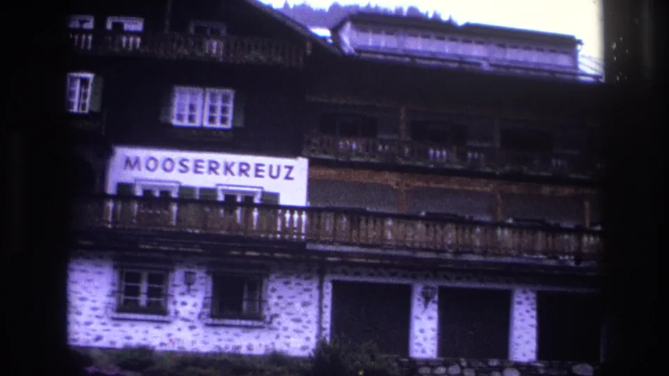 Upward pan showing Mooserkreuz sign against dark timber upper facade, stone walls and balcony railing below, conifer treeline behind, France, 1967.