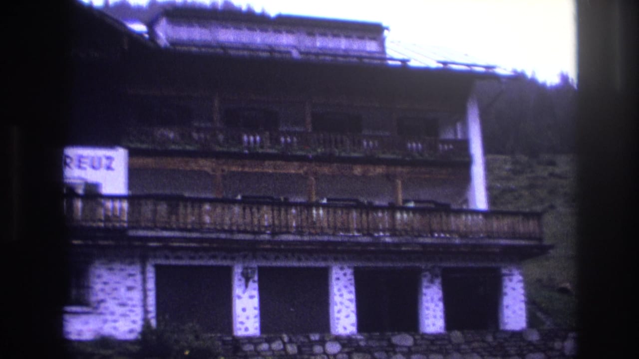 Multi-story alpine chalet with wooden balconies and stone foundation; partial sign text at frame edge; French Alps circa 1967, 8mm with purple color cast.