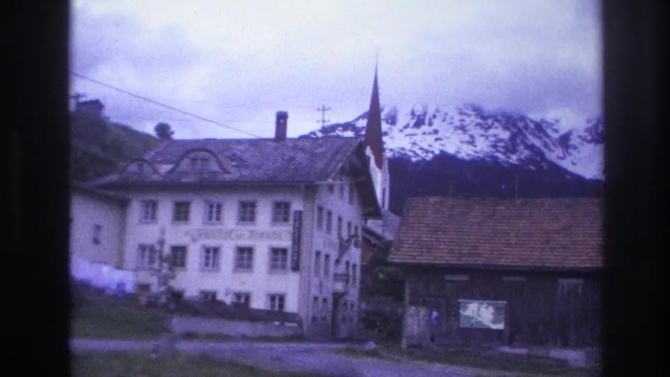 Alpine village street scene: white plaster inn, timber barn, church steeple with flag, snow-capped peaks; France circa 1967, 8mm home-movie footage.
