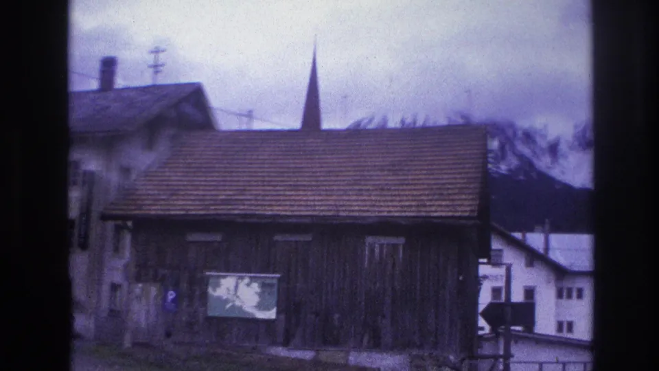 Timber barn and church steeple in an alpine village with snow-capped peaks; road sign and TV antenna visible; France circa 1967, 8mm home-movie footage.