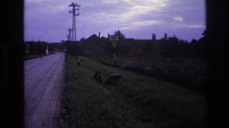 Rural road into an Alpine village circa 1967, with utility pylon, roadside signs, and a possible person seated on the grass verge, 8mm film.