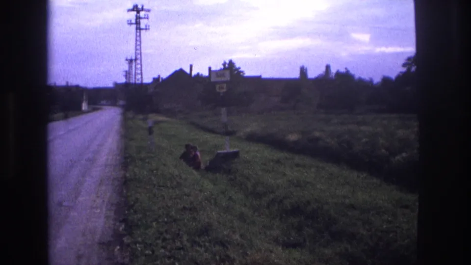 Alpine village approach road circa 1967, utility pylon and road signs visible, small figure on grass verge, 8mm home movie with blue-purple dye shift.