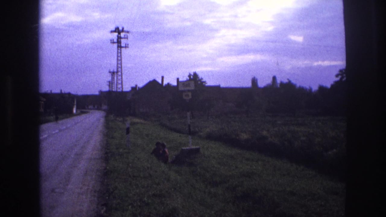 Rural country road with lattice electricity pylon, grassy verge, seated figure, and distant farm buildings, filmed from inside a moving vehicle.