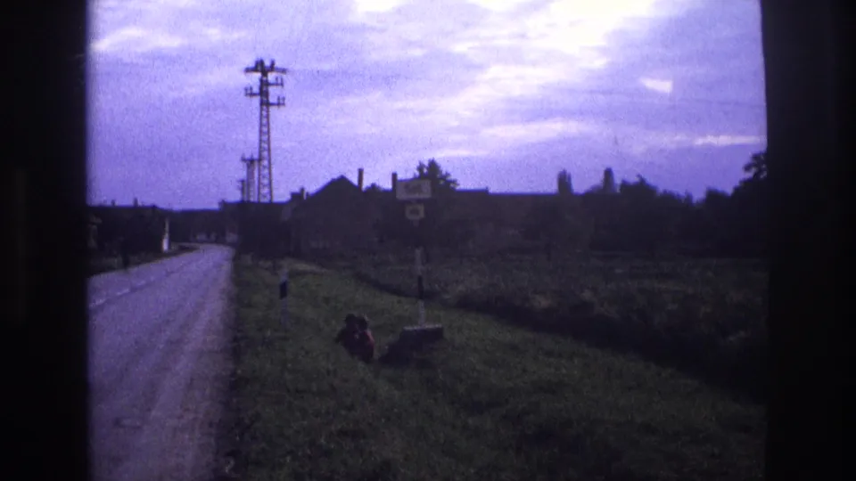 8mm home-movie frame of a rural road — per metadata, France 1967 — showing a lattice transmission tower, distant farm buildings, and a figure resting on the grass verge under a purple-blue sky.