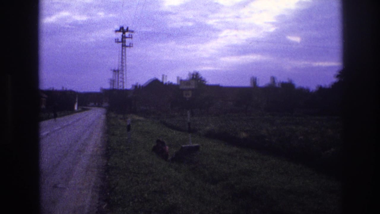 Panning 8mm frame of a rural road — per metadata, France 1967 — with a lattice pylon, a roadside sign, a seated figure on the verge, and a purple-blue overcast sky.
