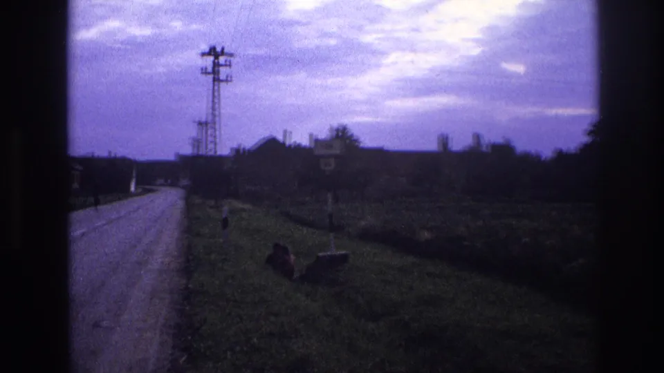 Panning 8mm frame of a rural road — per metadata, France 1967 — with a lattice pylon, a roadside sign, a seated figure on the verge, and a purple-blue overcast sky.