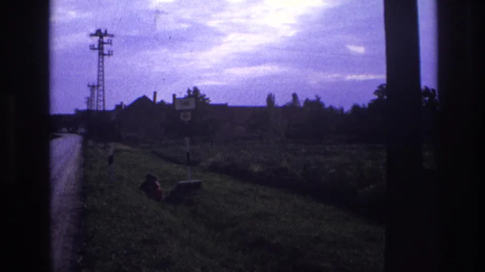 Mid-pan 8mm frame of grassy verge, roadside sign, lattice pylon, and distant farm buildings under a purple-blue sky; window-frame bar visible at centre; per metadata, France circa 1967.