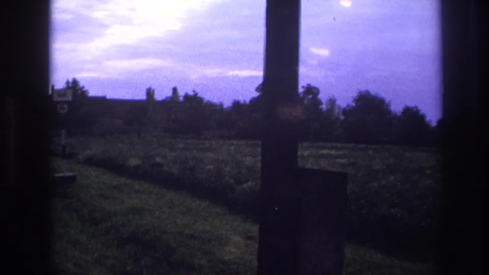 Final pan frame showing distant farm buildings, treeline, and a roadside sign under a purple-blue sky; a vehicle window post dominates the right edge; per metadata, France circa 1967.