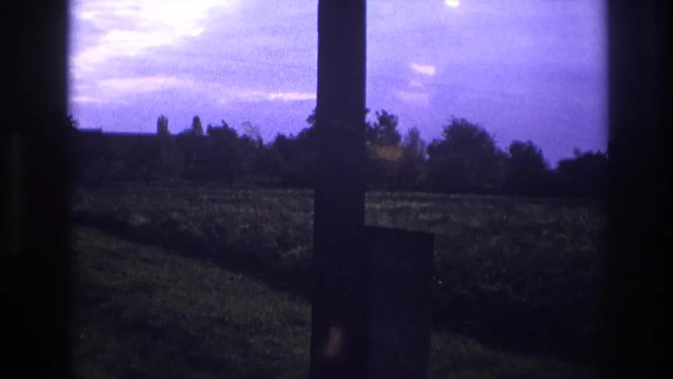 1967 France 8mm home movie — rural meadow seen through a window at dusk, deep purple sky with intense light flare and silhouetted trees.