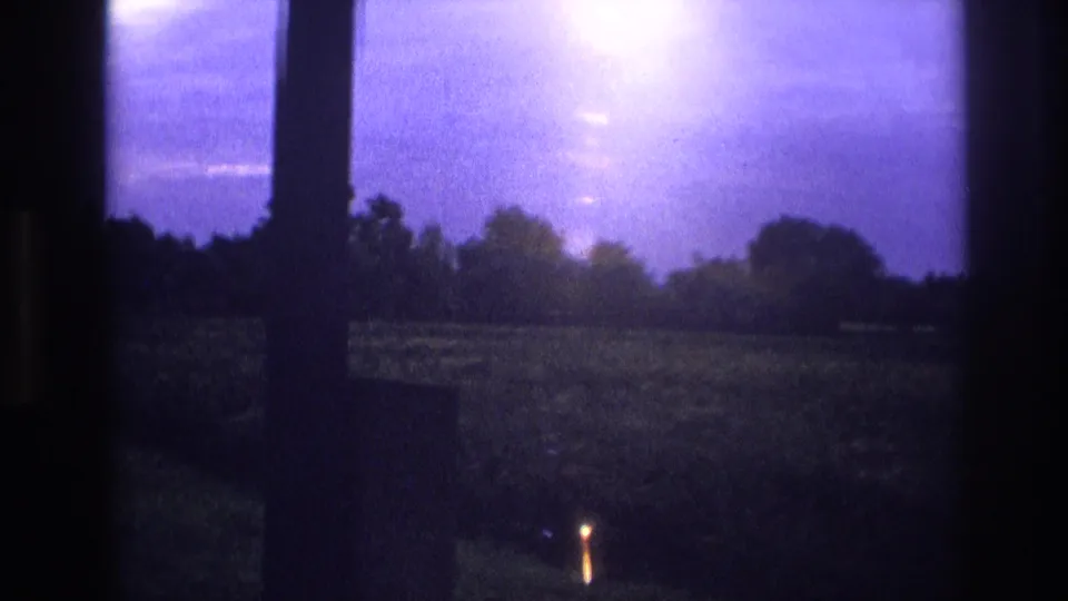 1967 France 8mm — rural field through window at dusk, prominent lens flare upper right, small warm light visible in the distant field.
