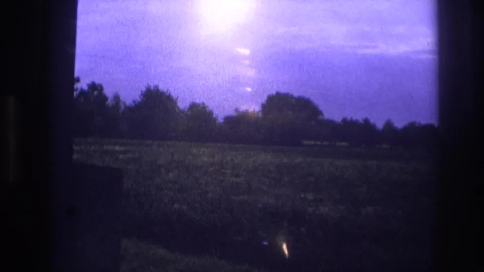1967 France 8mm — wider window view of rural meadow at dusk, intense light bloom upper left, treeline silhouetted against deep purple sky.