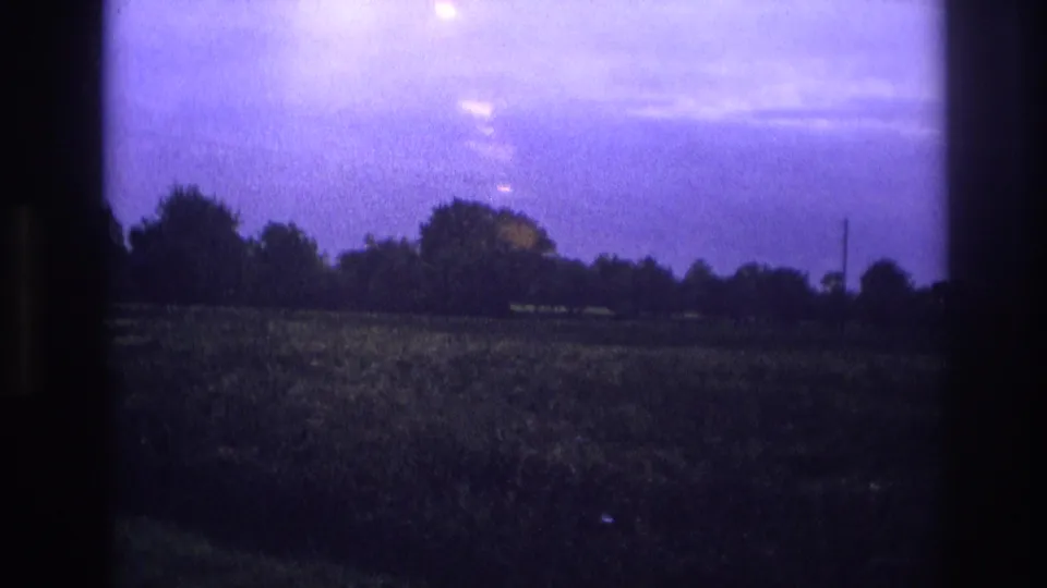 1967 France 8mm — rural landscape framed by window at dusk, light bloom upper right, treeline silhouette, warm distant point light at lower right.