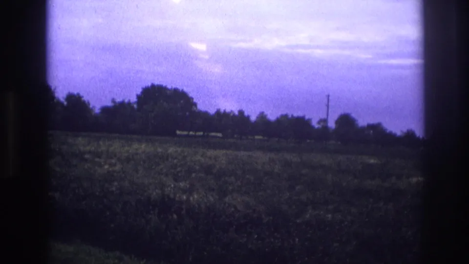 8mm home movie frame, rural France c.1967 — flat crop field beneath a purple-cast overcast sky with distant tree line and utility pole.